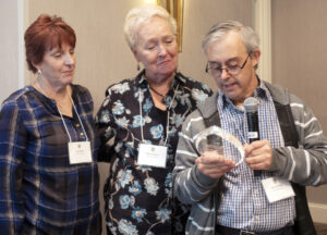 Chris Grandy is holding a microphone and reading what is on an award. Standing next to Chris is award recipient Nancy Richey. PADDC Council Member Lisa Butler is looking on.